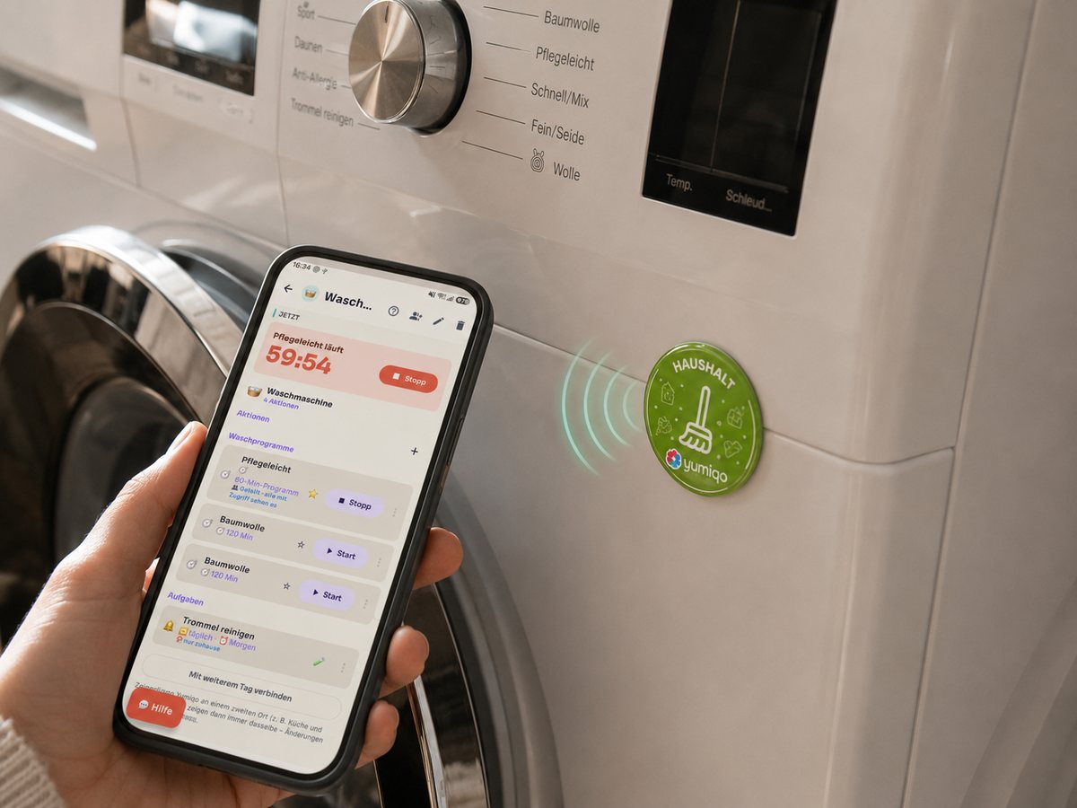 Eine Hand hält ein Smartphone mit geöffneter Yumiqo-App an einen grünen Haushalt-Yumiqo-Sticker auf der Waschmaschine – die App zeigt die laufende Wäsche und die nächsten Schritte.